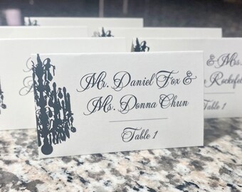 May include: White place card with black calligraphy text that reads "Mr. Daniel Fox & Ms. Donna Chun, Table 1". A black silhouette of a chandelier is in the upper left corner.