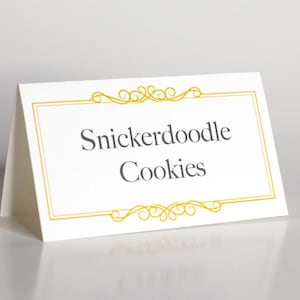 May include: A white card with a gold border and a decorative flourish design. The text "Snickerdoodle Cookies" is printed in gray.