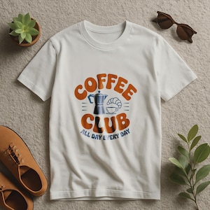 Puede incluir: Camiseta blanca con un gráfico que dice "Coffee Club" en naranja y azul. El gráfico incluye una cafetera, un cruasán y una taza de café. El texto "All Day Every Day" está debajo del texto del club.