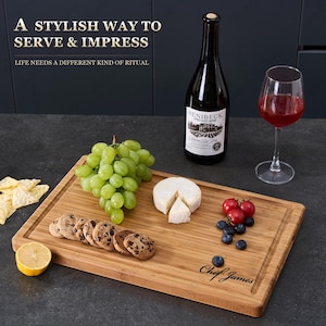 May include: A rectangular wooden serving board with a groove, holding grapes, cheese, cookies, blueberries, and tomatoes. A wine bottle and a glass of red wine are also present. The board reads "Chef James."