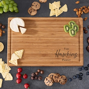 以下が含まれることがあります： 「The Hawkins Family」と刻まれた、縁がくぼんだ長方形の竹製まな板。チーズ、キウイ、クッキー、チップス、ブドウ、トマトなどのおやつに囲まれています。まな板は薄茶色です。