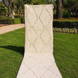 May include: A long, rectangular, off-white wool rug featuring a repeating diamond pattern outlined in thin, light brown lines. The rug has a thick, shaggy pile texture and is finished with a row of long, white knotted fringe along the bottom edge.