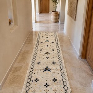 Puede incluir: Una alfombra de pasillo larga y rectangular de color crema con un borde de patrón de diamantes negros y un campo central decorado con pequeños motivos geométricos dispersos en negro, azul, amarillo y verde azulado, con flecos en los extremos.