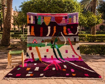 May include: A large, colorful rug with an abstract design, featuring a deep burgundy base and vibrant accents of pink, orange, green, and white. The rug is displayed outdoors, with a small wooden stool in the foreground.
