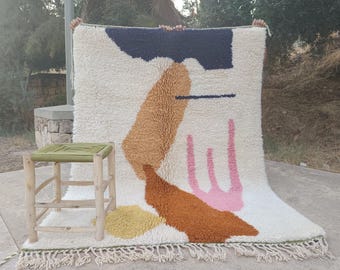 May include: A large, off-white rug with an abstract design featuring shapes in navy, brown, orange, pink, and yellow. A small, light green stool with a woven top sits to the left. The rug has a fringed edge.