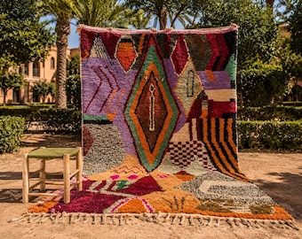 May include: A large, colorful Moroccan rug with geometric patterns in shades of purple, orange, green, and red. A small wooden stool with a green seat sits to the left of the rug. The rug has a fringed edge.