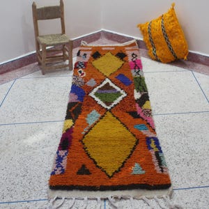 May include: An orange and multi-colored Moroccan rug with geometric patterns, including diamonds and triangles. The rug is rectangular with a white fringe. A small wooden chair and a yellow pillow are in the background.