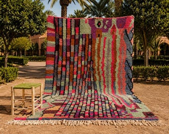 May include: A colorful, patterned rug with a fringed edge, displayed outdoors. The rug features a mix of vertical and horizontal rectangular designs in shades of red, pink, green, and brown. A small, square stool with a green woven top sits nearby.