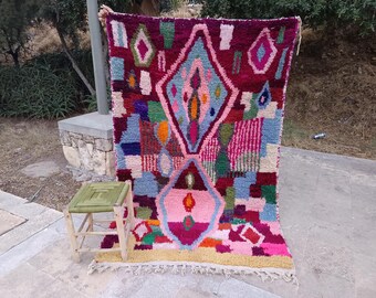 May include: A vibrant, hand-woven rug with a geometric design. The rug features a rich palette of colors, including burgundy, pink, blue, and green. The design includes diamond shapes and other abstract patterns. A small, green wooden stool sits nearby.