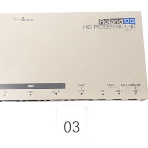 May include: A beige Roland DG MIDI Processing Unit with the text "MIDI PROCESSING UNIT" and "Roland DG" printed on the front. The unit has multiple input and output ports labelled "MIDI", "SYNC", "TAPE", and "METRONOME".