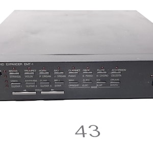 Può includere: Un Yamaha FM Sound Expander EMT-1 nero con una varietà di effetti sonori e controlli. L'unità ha un display digitale e una serie di pulsanti per selezionare diversi suoni, tra cui batteria, chitarre, corni e altro.