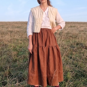 May include: A person in a cream quilted waistcoat and white blouse, with a long, tiered brown skirt. They are wearing burgundy socks and brown boots. The scene is a grassy field under a blue sky, suggesting a rustic or bohemian style, with a focus on natural fabrics.