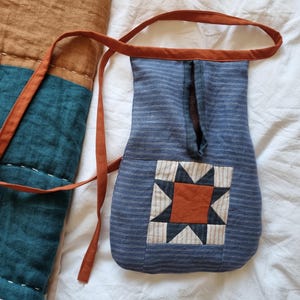 Peut inclure: Un sac en tissu bleu rayé avec une sangle marron et un écusson en forme d'étoile. Le sac est fait d'un tissu doux et texturé et a une forme rectangulaire. L'écusson en étoile est fait de tissu orange, blanc et bleu marine.