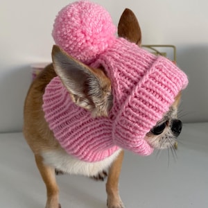 Peut inclure: Un snood tricoté rose clair avec une texture côtelée et un gros pompon assorti sur le dessus. L'accessoire comporte deux ouvertures pour les oreilles et est conçu pour s'ajuster autour du cou et de la tête d'un petit animal.