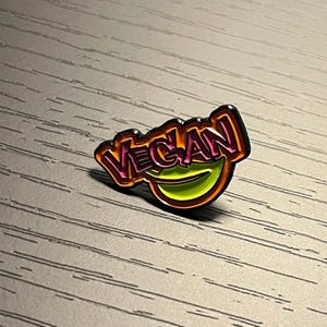Könnte beinhalten: Eine rosa und gelbe Emaille-Anstecknadel mit dem Wort "VEGAN" in einer fetten, abgerundeten Schrift. Die Anstecknadel hat einen schwarzen Umriss und ein grünes, gelbes und orangefarbenes Design in der Mitte.