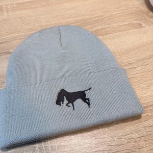 Puede incluir: Un gorro azul claro con una silueta negra de un perro. El gorro está hecho de tejido de punto acanalado. La silueta del perro está bordada en la parte delantera del gorro.