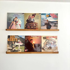 May include: Two wooden shelves with a collection of vintage vinyl records. The records have colorful covers with German text and images of people and birds. The shelves are mounted on a white wall.