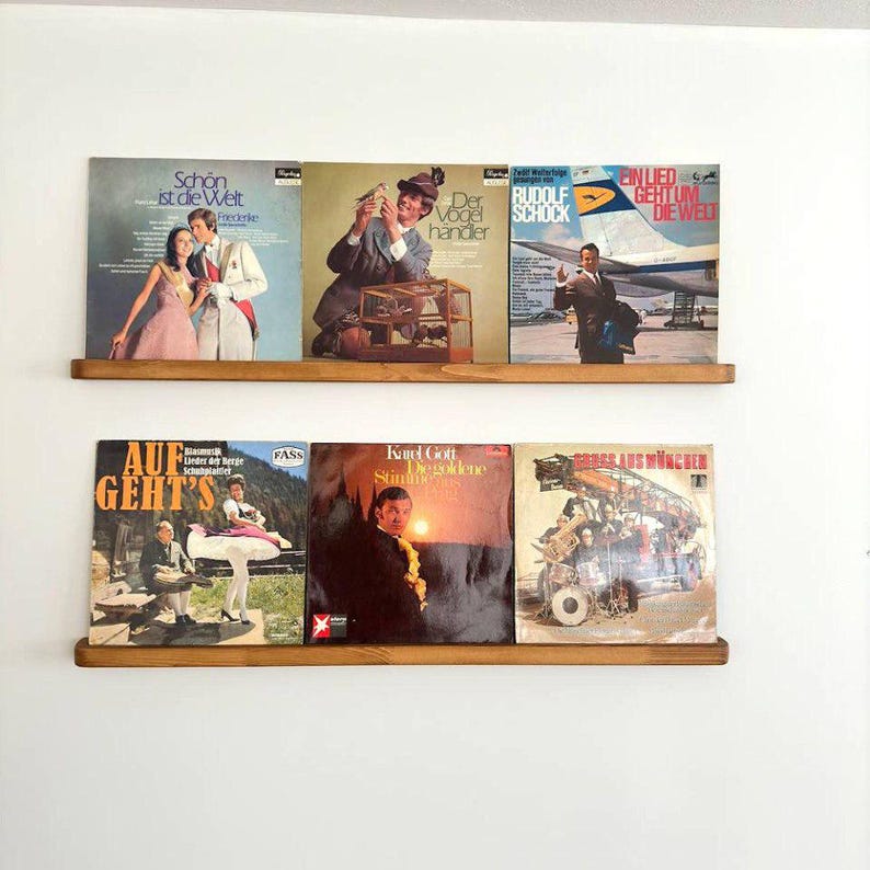 May include: Two wooden shelves with a collection of vintage vinyl records. The records have colorful covers with German text and images of people and birds. The shelves are mounted on a white wall.