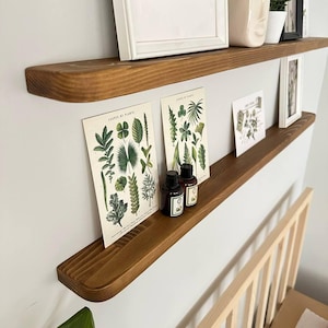 Può includere: Due mensole in legno fissate a una parete bianca, che espongono arte incorniciata, stampe botaniche, piccole piante in vaso e bottiglie decorative. Le mensole sono tinte di un marrone caldo, creando un contrasto con la parete bianca. Le stampe botaniche presentano illustrazioni di foglie verdi.