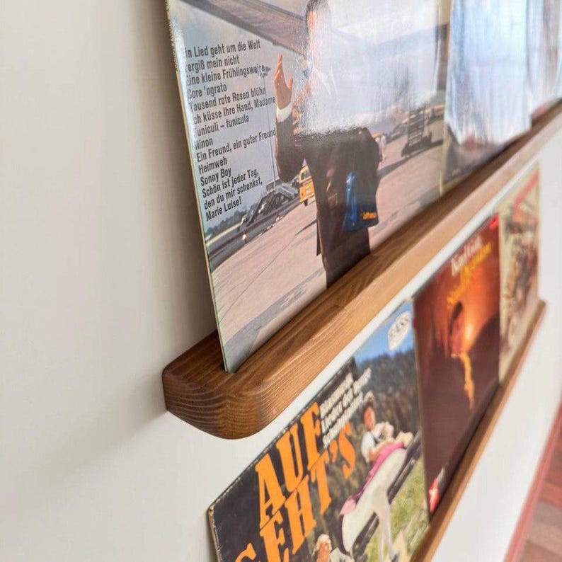 May include: Two wooden shelves mounted on a white wall, displaying vinyl records. The shelves are stained a dark brown color and have a rounded edge. The top shelf has a record with German text, while the bottom shelf has a record with the text "AUF GEHT'S".