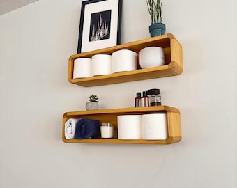 May include: Two wooden shelves with rounded edges, mounted on a white wall. The top shelf has three rolls of toilet paper and a small potted plant. The bottom shelf has two rolls of toilet paper, a blue hand towel, a small candle, and several small bottles.