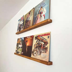 May include: Two wooden shelves mounted on a white wall, displaying a collection of vintage vinyl records. The shelves are made of natural wood with a rustic finish. The records feature a variety of genres and artists, including a record with a picture of a car.