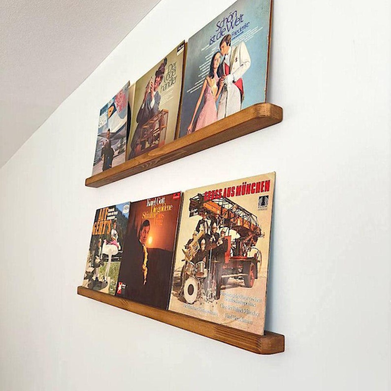 May include: Two wooden shelves mounted on a white wall, displaying a collection of vintage vinyl records. The shelves are made of natural wood with a rustic finish. The records feature a variety of genres and artists, including a record with a picture of a car.