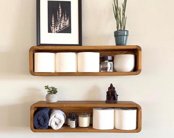 May include: Two wooden shelves with rounded edges, mounted on a white wall. The top shelf has three rolls of toilet paper, a small bottle of perfume, and a white ceramic cup. The bottom shelf has two rolls of toilet paper, two towels, a small bottle of lotion, a candle, and a small wooden figurine.