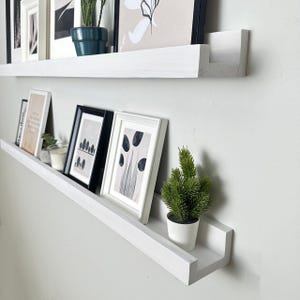 May include: Two white floating shelves mounted on a light gray wall. The shelves display framed art, small potted plants, and a candle. The art includes black and white prints and a framed photograph.