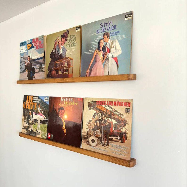 May include: Two wooden shelves with vinyl records on them. The shelves are mounted on a white wall. The records have various titles and artists, including "Der Vogelh&auml;ndler", "Sch&ouml;n ist die Welt", "Auf Geht's", "Karl Gott", and "Express aus M&uuml;nchen".