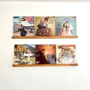 May include: A wooden shelf with eight vinyl records displayed. The records have various titles and artists, including "Rudolf Schock", "Ein Lied Geht Um Die Welt", "Der Vogelh&auml;ndler", "Sch&ouml;n Ist Die Welt", "Auf Geht's", "Karel Gott", and "Gross Aus M&uuml;nchen".