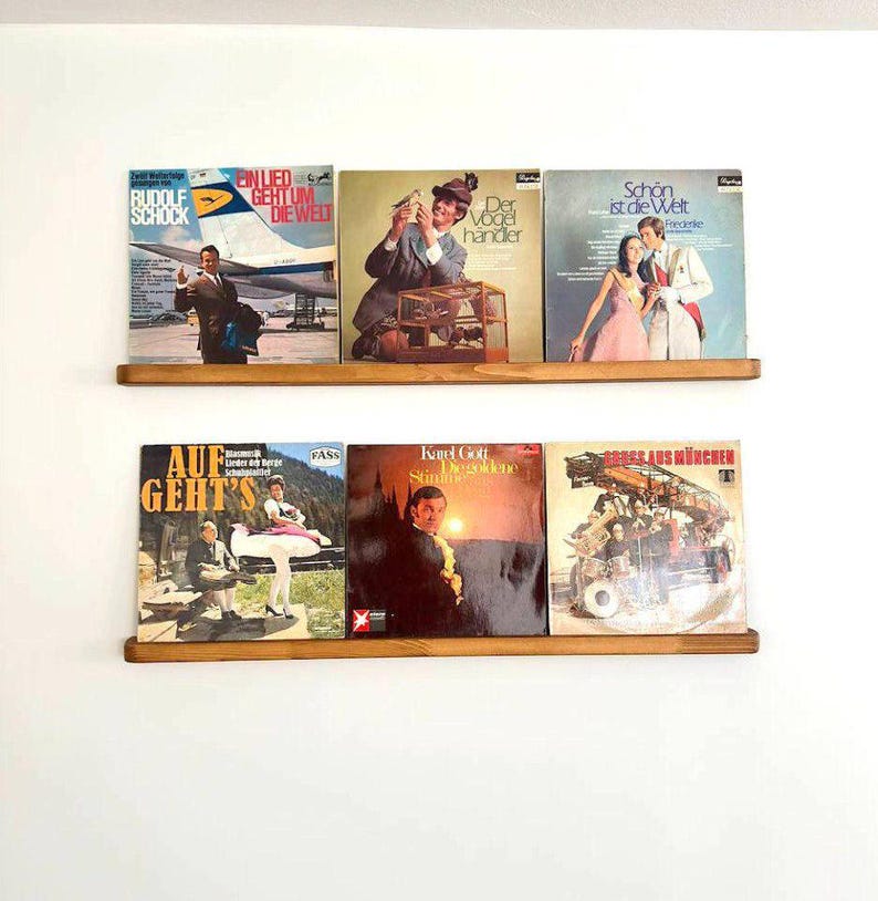 May include: A wooden shelf with eight vinyl records displayed. The records have various titles and artists, including "Rudolf Schock", "Ein Lied Geht Um Die Welt", "Der Vogelh&auml;ndler", "Sch&ouml;n Ist Die Welt", "Auf Geht's", "Karel Gott", and "Gross Aus M&uuml;nchen".
