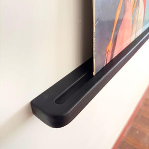 May include: Black wooden wall shelf with a record leaning against it. The shelf has a groove to hold the record in place.