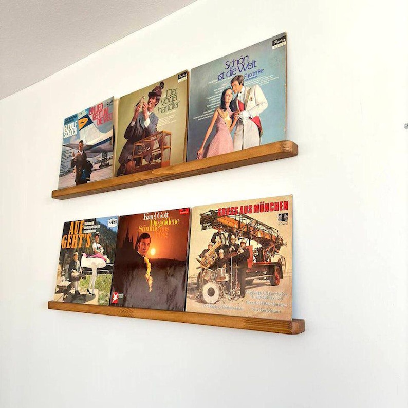 May include: Two wooden shelves mounted on a white wall, displaying a collection of vintage vinyl records. The records feature various artists and album covers, including a record with the text "Schon ist die Welt" and another with the text "Der Vogelh&auml;ndler".