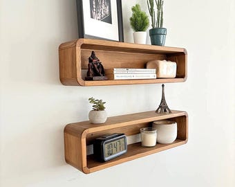 May include: Two wooden wall shelves with rounded edges. The top shelf has a framed print, a small statue, and books. The bottom shelf has a small plant, a clock, a candle, and a small Eiffel Tower statue.