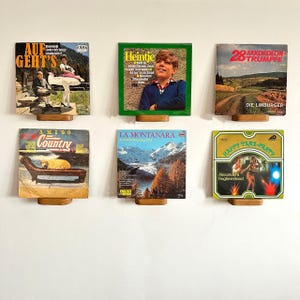 May include: Six vintage vinyl record album covers displayed on wooden stands. The album covers feature various designs, including landscapes, portraits, and text. The albums are mounted on small wooden stands, creating a unique wall display.