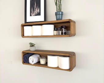 May include: Two wooden shelves with rounded edges, mounted on a white wall. The top shelf has three rolls of toilet paper, a small potted plant, and several glass bottles. The bottom shelf has a rolled towel, a candle, and two rolls of toilet paper.