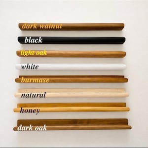 Puede incluir: Una selección de estantes de madera en varios colores, incluyendo nogal oscuro, negro, roble claro, blanco, birmano, natural, miel y roble oscuro. Cada estante tiene un borde frontal redondeado y está etiquetado con su nombre de color.