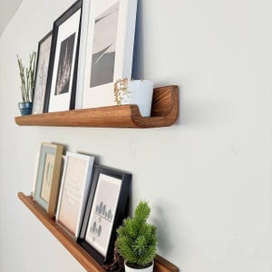 May include: Two wooden shelves with a curved edge, mounted on a white wall. The shelves are decorated with framed pictures and potted plants.