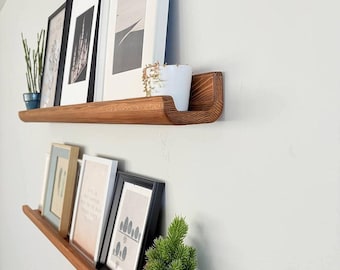 May include: Two wooden shelves with a curved edge, mounted on a white wall. The shelves are decorated with framed pictures and potted plants.