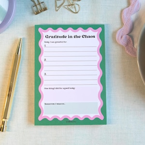 May include: A notepad with the text "Gratitude in the Chaos" in green and pink sits on a light green scalloped placemat. A gold pen, a cup of tea, and a plate with a croissant and berries are also visible.