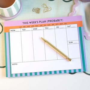 Pode incluir: Um planejador semanal com um design colorido. O planejador apresenta um interior branco com seções para cada dia da semana e o texto "THIS WEEK'S PLAN (PROBABLY)". Uma caneta dourada repousa sobre o planejador. É cercado por uma caneca, um prato e itens decorativos.