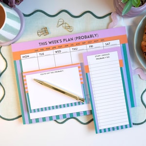 Könnte beinhalten: Ein farbenfroher Wochenplaner mit dem Text "THIS WEEK'S PLAN (PROBABLY)". Der Planer enthält Abschnitte für jeden Wochentag, eine To-Do-Liste und einen Notizblock. Ein goldener Stift liegt auf dem Notizblock. Ein Croissant und Beeren liegen auf einem Teller.