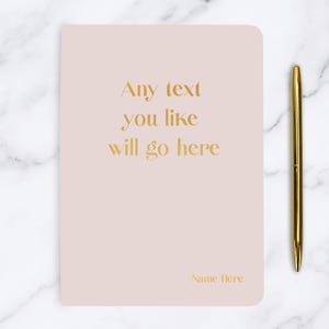 Peut inclure: Un carnet rose pâle avec un lettrage doré qui dit "Any text you like will go here" et "Name Here" en bas.