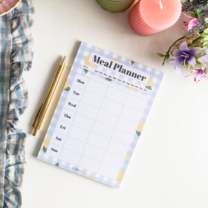 Op de afbeelding: Een blauw en wit geruit notitieboekje met een citroendesign. Het notitieboekje heeft de kop "Meal Planner" en een kalender met de maanden van het jaar. Het notitieboekje ligt op een wit oppervlak met een pen.