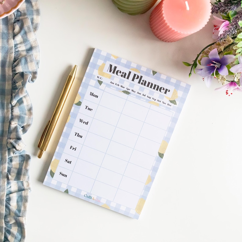 Op de afbeelding: Een blauw en wit geruit notitieboekje met een citroendesign. Het notitieboekje heeft de kop "Meal Planner" en een kalender met de maanden van het jaar. Het notitieboekje ligt op een wit oppervlak met een pen.