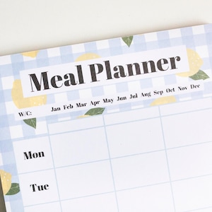 Op de afbeelding: Een witte maaltijdplanner met een blauw-wit geruit patroon. De planner heeft een citroenmotief en de woorden "Meal Planner" bovenaan. De planner is verdeeld in maanden en dagen van de week.