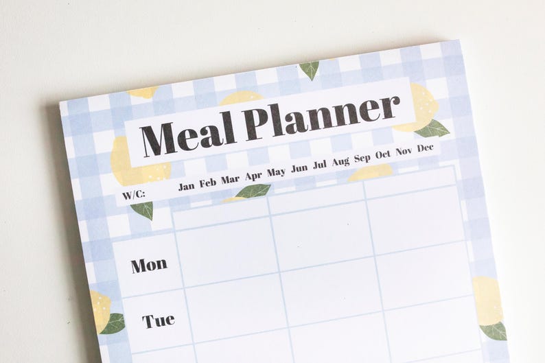 Op de afbeelding: Een witte maaltijdplanner met een blauw-wit geruit patroon. De planner heeft een citroenmotief en de woorden "Meal Planner" bovenaan. De planner is verdeeld in maanden en dagen van de week.