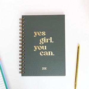 May include: A dark teal spiral-bound notebook with gold lettering that reads "yes girl, you can." The name "ZOE" is printed below the text. A gold pen and a notepad are also visible.