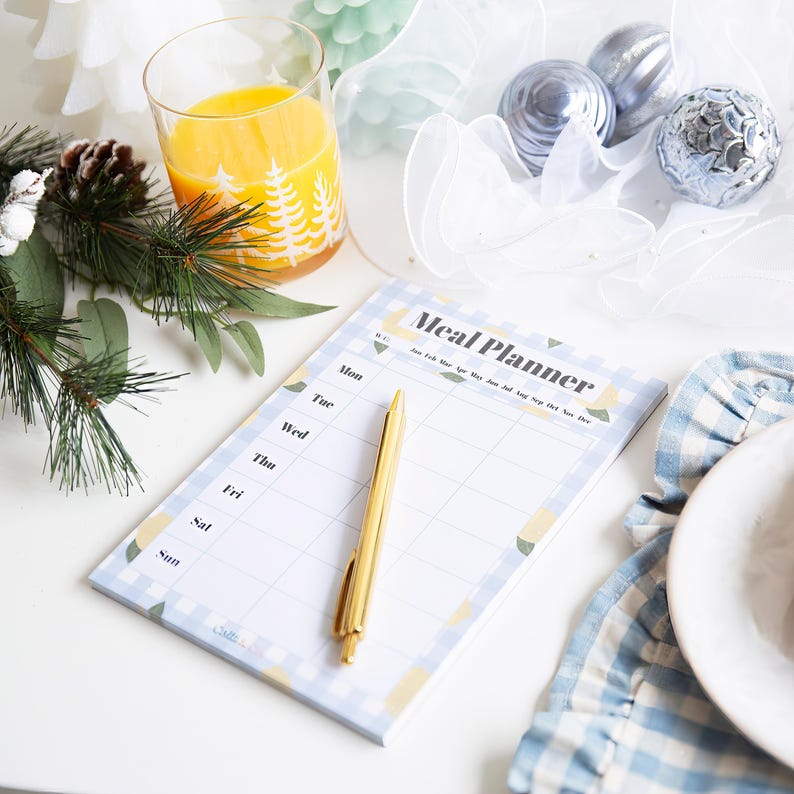 Op de afbeelding: Een blauwe en witte maaltijdplanner notitieboekje met een citroendesign. Het notitieboekje heeft een gouden pen erop en de woorden "Meal Planner" staan bovenaan het notitieboekje.